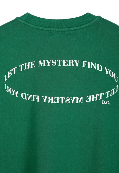 Chemise verte avec un texte blanc incurvé indiquant "LET THE MYSTERY FIND YOU" répété deux fois, avec les initiales "B.C." en bas à droite.