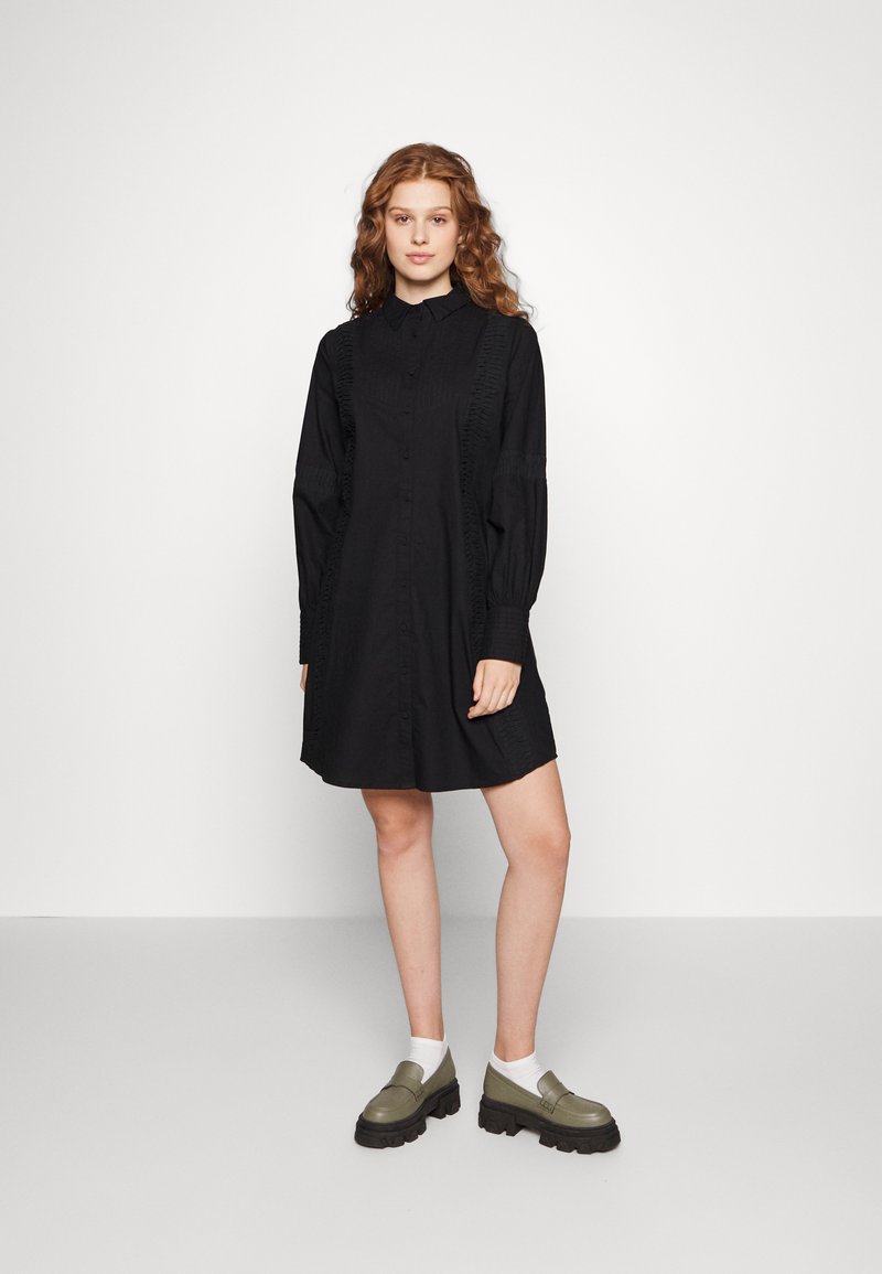 Vestido preto de botões com mangas longas e detalhes texturizados. Apresenta um corte descontraído e termina acima do joelho. Combinado com sapatos verdes robustos.