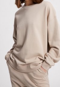Felpa beige con vestibilità morbida, scollo rotondo, spalle scese e polsini a costine. Tessuto morbido con texture minima, abbinata a pantaloni coordinati.