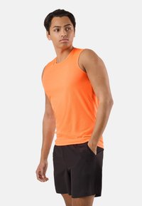 Jeune homme portant un débardeur de sport orange et un short noir, debout avec une main dans la poche, regardant sur le côté.