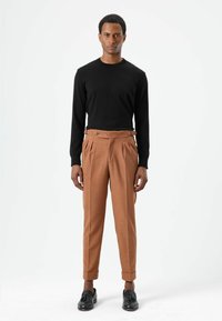 Zwart shirt met lange mouwen gecombineerd met beige geplooide broek. De broek heeft een knoopdetail en is aan de onderkant omgeslagen. Zwarte schoenen maken de look af.