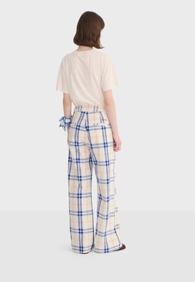 Personne de dos portant une chemise beige à manches courtes et un pantalon large crème avec un motif à carreaux bleu et orange, debout sur un fond blanc.