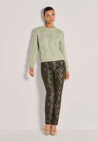 Frau trägt einen hellgrünen Kabelstrickpullover, Schlangenprint-Hosen und beige spitze Absätze und steht vor einer schlichten hellen Wand.