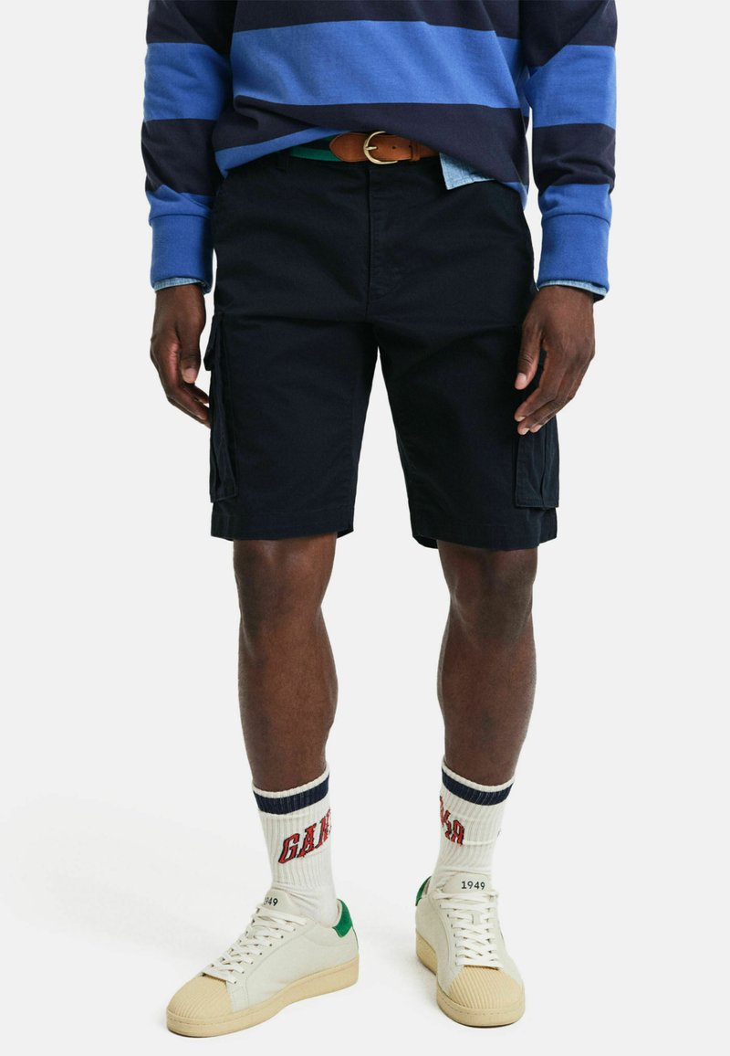 Pantalones cortos de carga en color navy con bolsillos laterales, fabricados en algodón, que incluyen un cinturón con una hebilla marrón. Se llevan con un sudadera a rayas en azul y negro.