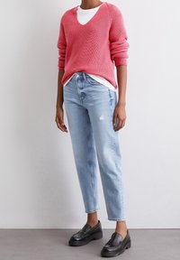 Un pull en V tricoté rose superposé à une chemise blanche, associé à un jean droit déchiré bleu clair et des mocassins noirs sans lacets.
