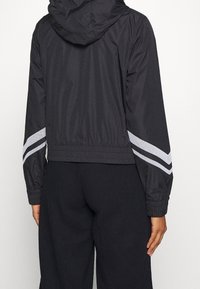 Mustat tuulitakki hupulla, jossa on hopeiset heijastavat raidat hihoissa, joustavat hihansuut ja lyhyt muotoilu. Sileä pinta.