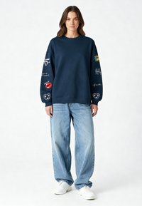 Felpa blu navy con maniche lunghe, caratterizzata da diverse stampe colorate e ricami. Abbinata a jeans baggy blu chiaro e sneakers bianche.