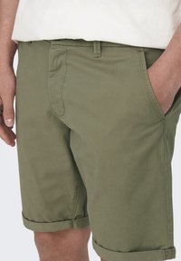 Shorts en coton vert olive avec des ourlets retroussés, des poches avant et une texture lisse. Comprend des passants de ceinture et des détails de couture discrets.