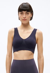 Moteris su trumpais tamsiais plaukais, dėvinti tamsiai violetinę be siūlių sportinę liemenėlę ir aukšto liemens aukštos juosmens tamprius, stovinti tiesiai priešais paprastą baltą foną.