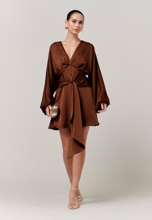 Kvinde i en brun satin indpakningskjole med lange puffede ærmer, der holder en lille guldtaske, iført gyldne øreringe og sandaler med stropper.