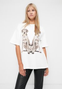 Witte katoenen t-shirt met een grafische illustratie van twee zittende honden met een gouden kettingaccent. Korte mouwen, losse pasvorm, eenvoudig ontwerp.