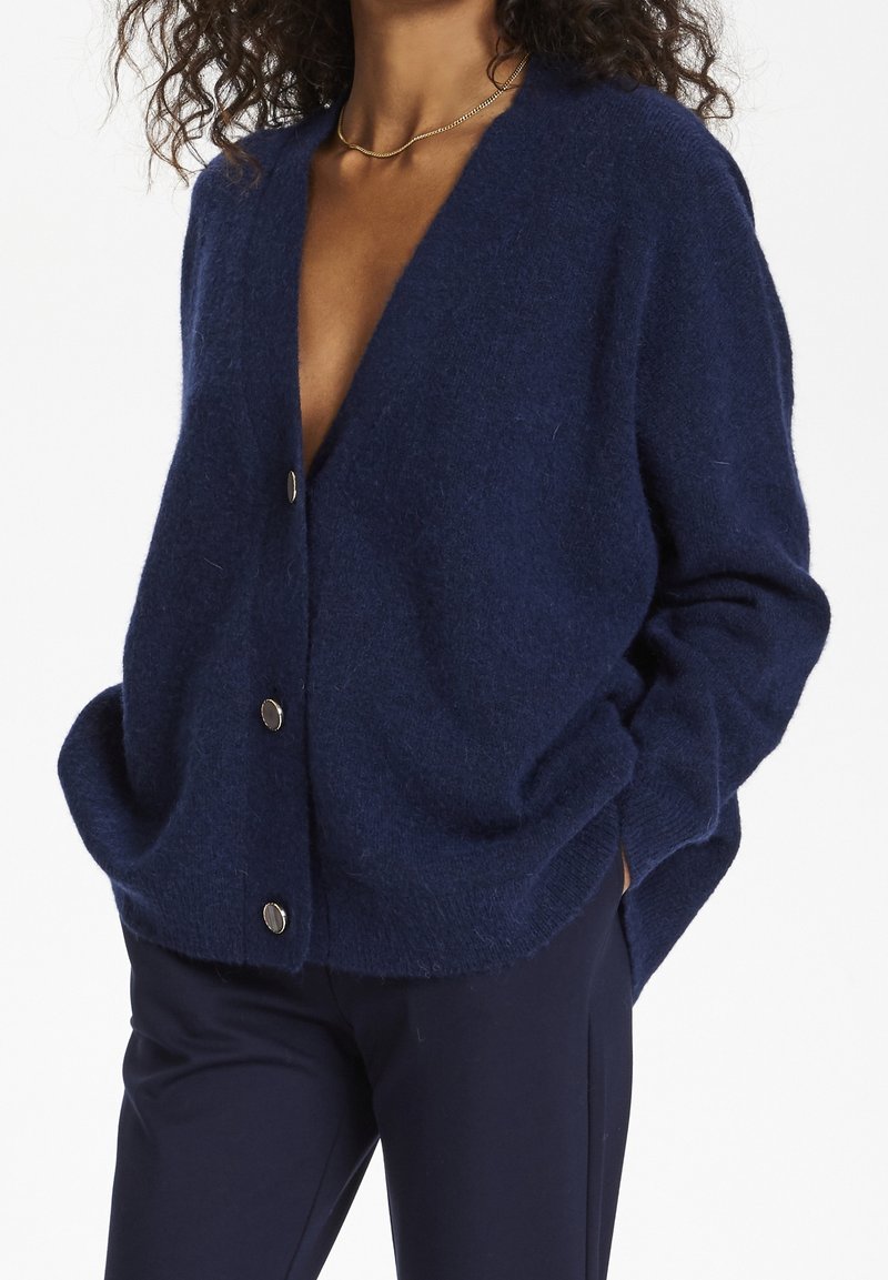 Marinblå cardigan gjord av mjukt tyg, med djup V-ringning, stora sidofickor och metallknappar framtill. Avslappnad passform.