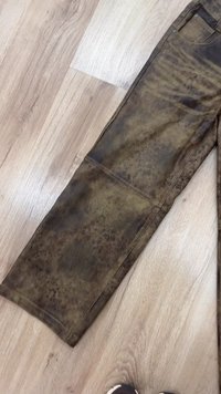 Rjave hlače iz umetnega usnja z teksturirano površino, s ravenim krojem in vidnimi šivi. Tkanina ima madežast vzorec.