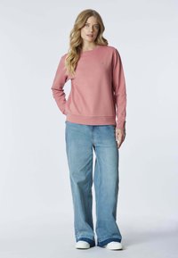 Felpa rosa con maniche lunghe, scollo rotondo e polsini e orlo a coste. Abbinata a jeans a gamba larga di colore azzurro chiaro e scarpe da ginnastica bianche.