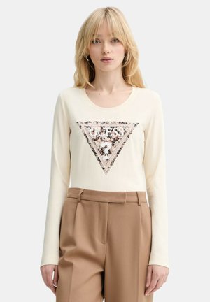 Donna bionda che indossa una maglia crema a maniche lunghe con un logo triangolare Guess di paillettes e pantaloni color cammello a vita alta, in piedi.