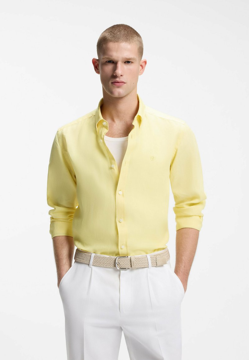 Jeune homme aux cheveux courts portant une chemise jaune boutonnée sur un sous-t-shirt blanc, un pantalon blanc et une ceinture tissée beige, les mains dans les poches.