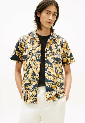 Junger Mann trägt ein schwarzes Hemd und ein offenes, kurzärmliges Hemd mit gelbem und orangefarbenem tropischem Blattmuster, kombiniert mit weißen Hosen.