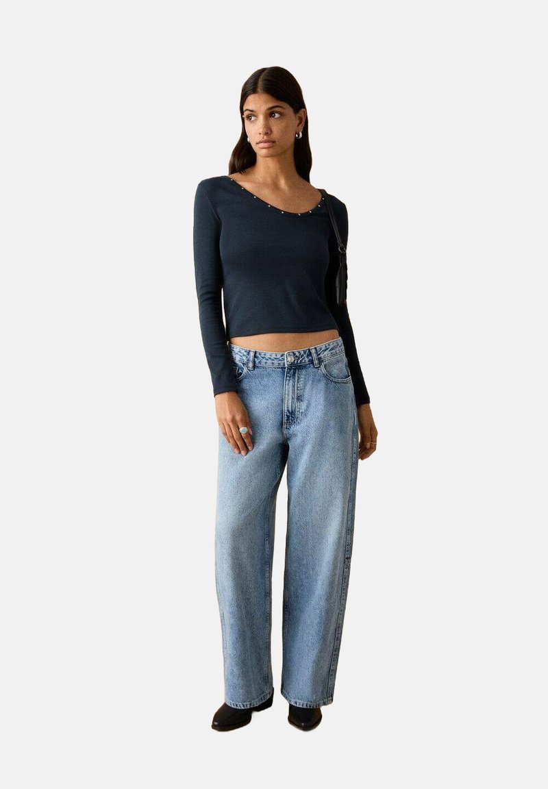 Marineblaues Langarmshirt mit U-Boot-Ausschnitt und kleinen Zierstollen, kombiniert mit locker sitzenden hellblauen Jeans und schwarzen Schuhen mit Absatz.