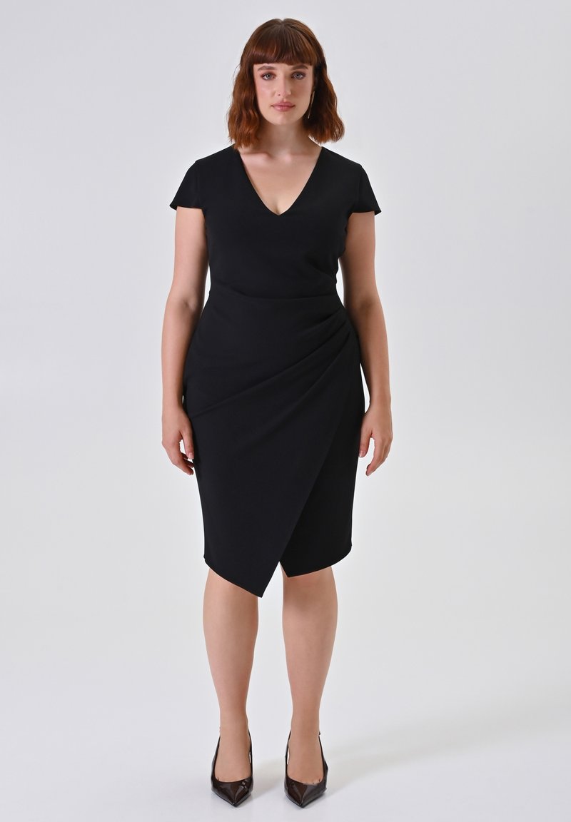 Frau mit schulterlangem kastanienbraunem Haar, die ein schwarzes V-Ausschnitt-Wickelkleid und schwarze spitze Absätze vor einem schlichten hellen Hintergrund trägt.