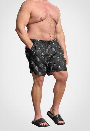 Mężczyzna bez koszulki, ubrany w czarne kąpielówki z wzorem czaszki i piszczeli oraz czarne klapki Nike, stoi na jednolitym białym tle.