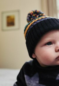 Strickmütze mit einem mehrfarbigen Pompon, aus dunklem Stoff und gestreiften Akzenten in Grün, Orange und Lila. Gesicht teilweise sichtbar.
