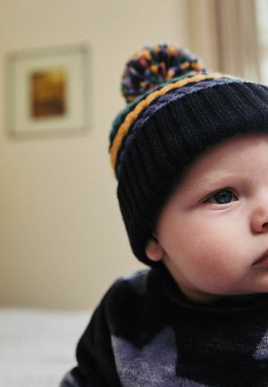 Bonnet en tricot avec un pompon multicolore, en tissu foncé et avec des accents rayés en vert, orange et violet. Visage partiellement visible.