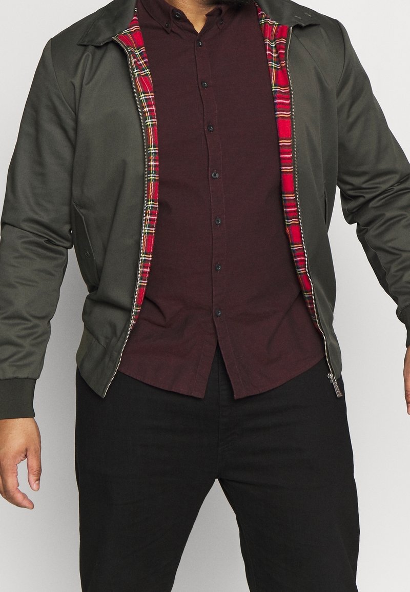 Homme portant une veste vert foncé avec une doublure intérieure à carreaux rouges sur une chemise bordeaux foncé boutonnée et un pantalon noir.