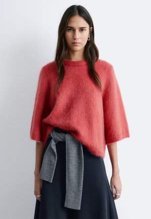 Vrouw met lang donker haar, gekleed in een loszittende, roodachtige, pluizige trui met driekwart mouwen en een grijze trui met strik om haar middel.