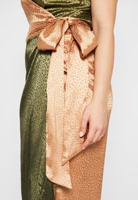 Robe portefeuille en satin avec un motif léopard dans des nuances de vert et de bronze, dotée d'un lien à l'avant et d'un tissu texturé. Main montrée reposant sur la robe.
