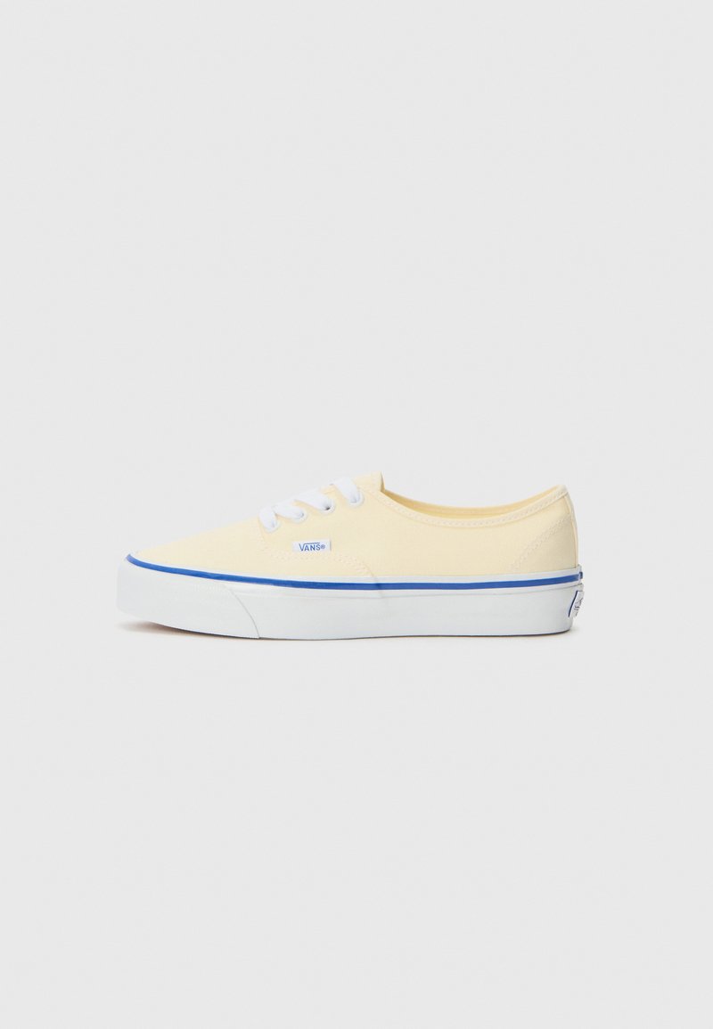 Canvas sneakers in pale yellow with white laces, blue stripe along the sole, flat rubber outsole, and Vans logo. Low-top design.