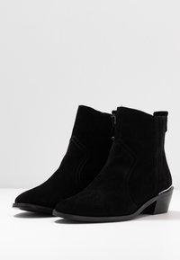 Para czarnych zamszowych botków za kostkę z niskimi, prostokątnymi obcasami i bocznymi zamkami na białej powierzchni i jasnoszarym tle.