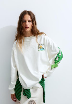 Ung kvinde med langt bølget hår iført en hvid oversized sweatshirt med grønt flamedesign på ærmet og bjerg-kyst grafik på brystet.