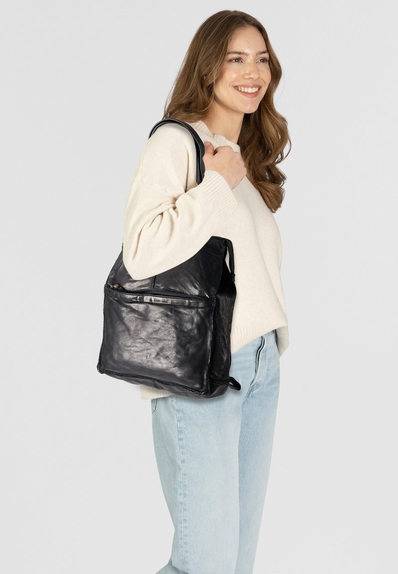 Femme souriante portant un pull léger et un jean, tenant un grand sac à bandoulière en cuir noir avec une poche zippée à l'avant, sur fond uni.