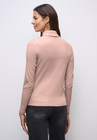 Lichtroze top met lange mouwen en een hoge hals, met een gladde textuur en een slanke pasvorm, gecombineerd met donkere grijze spijkerbroeken. Achteraanzicht toont de eenvoud van het ontwerp.