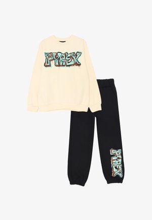 Felpa crema con grafica colorata "PYREX" sul davanti, abbinata a pantaloni della tuta neri con lo stesso logo sulla gamba. Tessuto in misto cotone.