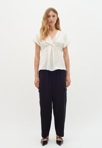 Blusa annodata di colore crema con maniche corte; abbinata a pantaloni cargo neri, larghi e scarpe basse nere a punta.