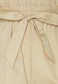 Beige tygmidja med en stor rosettknutdetalj fram. Slät textur med samlade stygn för elastisk passform.