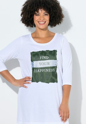 Donna sorridente con capelli ricci che indossa una camicia bianca con grafica verde e striscia argento con la scritta "FIND YOUR HAPPINESS", posa con la mano sul fianco.