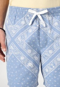Lyseblå svømmebukser laget av stoff med et hvitt mønster, med elastisk midje og hvit snor for feste.