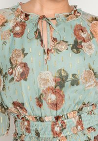 Bluse mit floralem Muster in Mintgrün, mit einem Rüschenkragen, Zugband und goldenen Akzentmustern. Verfügt über rosafarbene und burgunderfarbene Blumenprints.