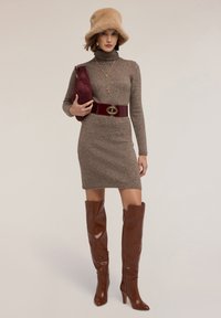 Braunes Strickkleid mit Rollkragen, einem burgunderfarbenen Gürtel mit goldenen Details, braunen Lederstiefeln bis zum Oberschenkel und einem weichen beigen Pelzhut.