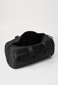Bolsa de deporte negra hecha de tela duradera, con dos asas y una parte superior con cremallera. Presenta una marca sutil y un diseño minimalista.