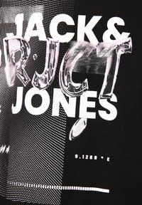 Sort t-shirt med fed, hvid tekst med en struktureret, blank effekt. Inkluderer grafiske elementer og et prikket mønster for kontrast.