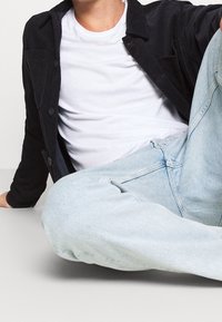 Mörk kordjacka med knappar över en vit t-shirt, tillsammans med ljusa denimjeans. Släta texturer och avslappnad passform synliga.