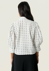 Blouse blanche avec un motif de nœuds noirs, manches bouffantes et col. Tissu texturé avec un ourlet droit et des poignets à boutons.