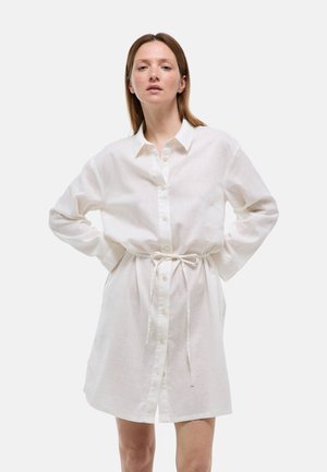 Femme aux cheveux raides portant une robe-chemise blanche à manches longues boutonnée, nouée à la taille, debout les mains sur les hanches devant un fond uni.