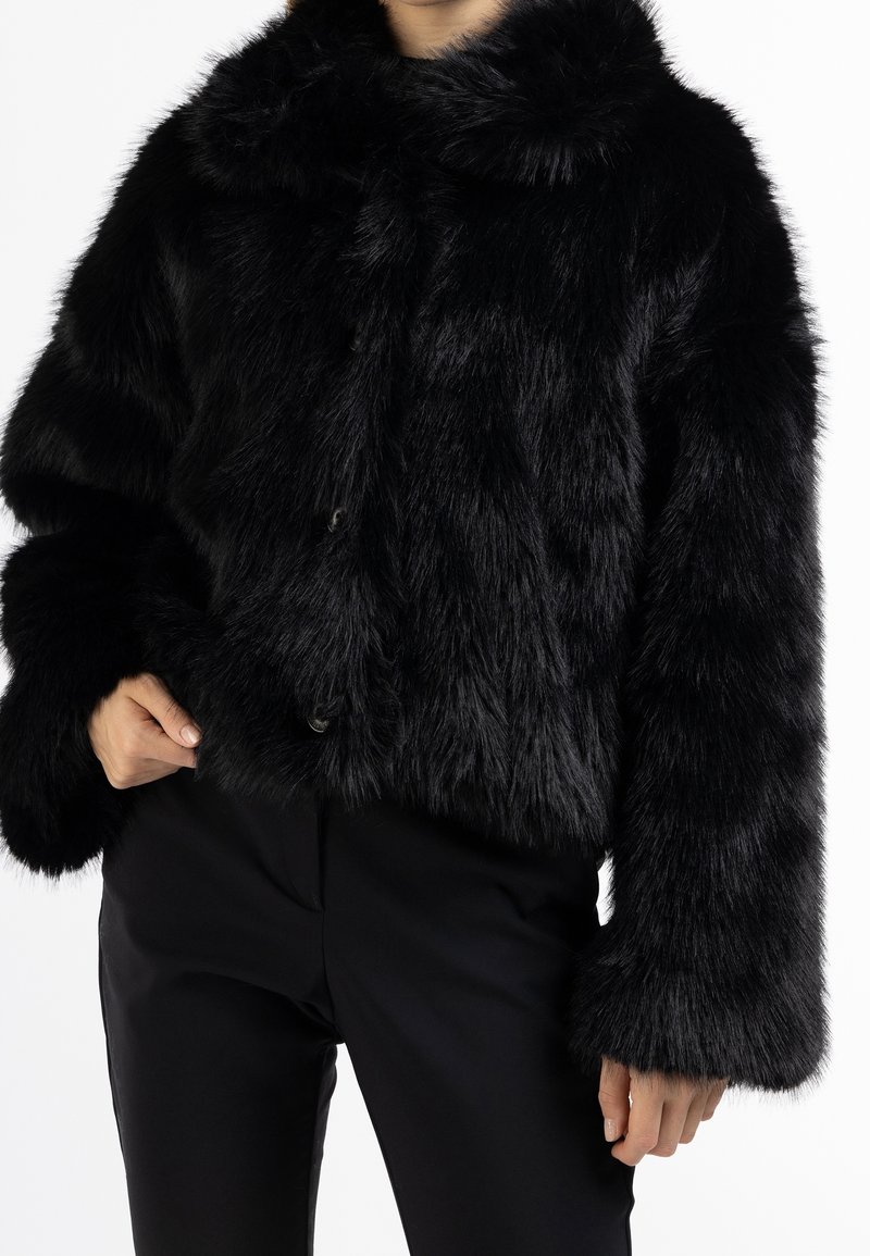 Personne portant une veste en fausse fourrure noire avec des manches larges et un col haut, associée à un pantalon noir, sur un fond blanc uni.