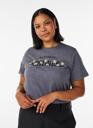 Donna con capelli scuri fino alle spalle che indossa una maglietta grigia con "All we need is kindness" e margherite, in piedi con le mani leggermente intrecciate.