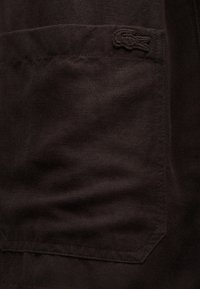 Tissu brun foncé avec une poche cousue montrant un petit logo de crocodile brodé près du bord supérieur.