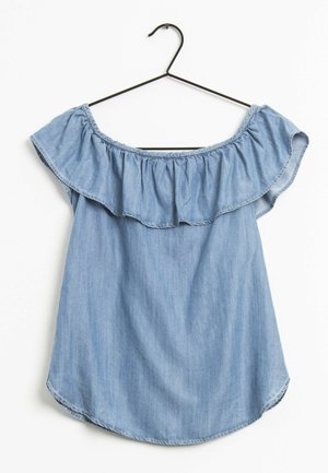 Blouse en jean bleu clair à épaules dénudées avec un col à volants suspendue sur un cintre en fil de fer noir sur un fond blanc.